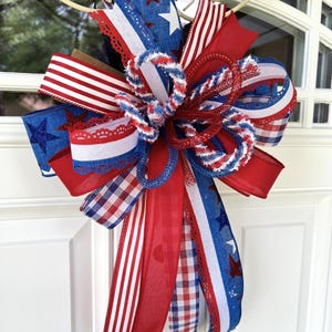 Op de afbeelding: Een decoratieve strik met een patriottisch thema in rood, wit en blauw. Het ontwerp bevat diverse patronen zoals sterren, strepen en gingham-ruitjes, gecombineerd met gestructureerd gaas en stoffen linten met een kanten rand.