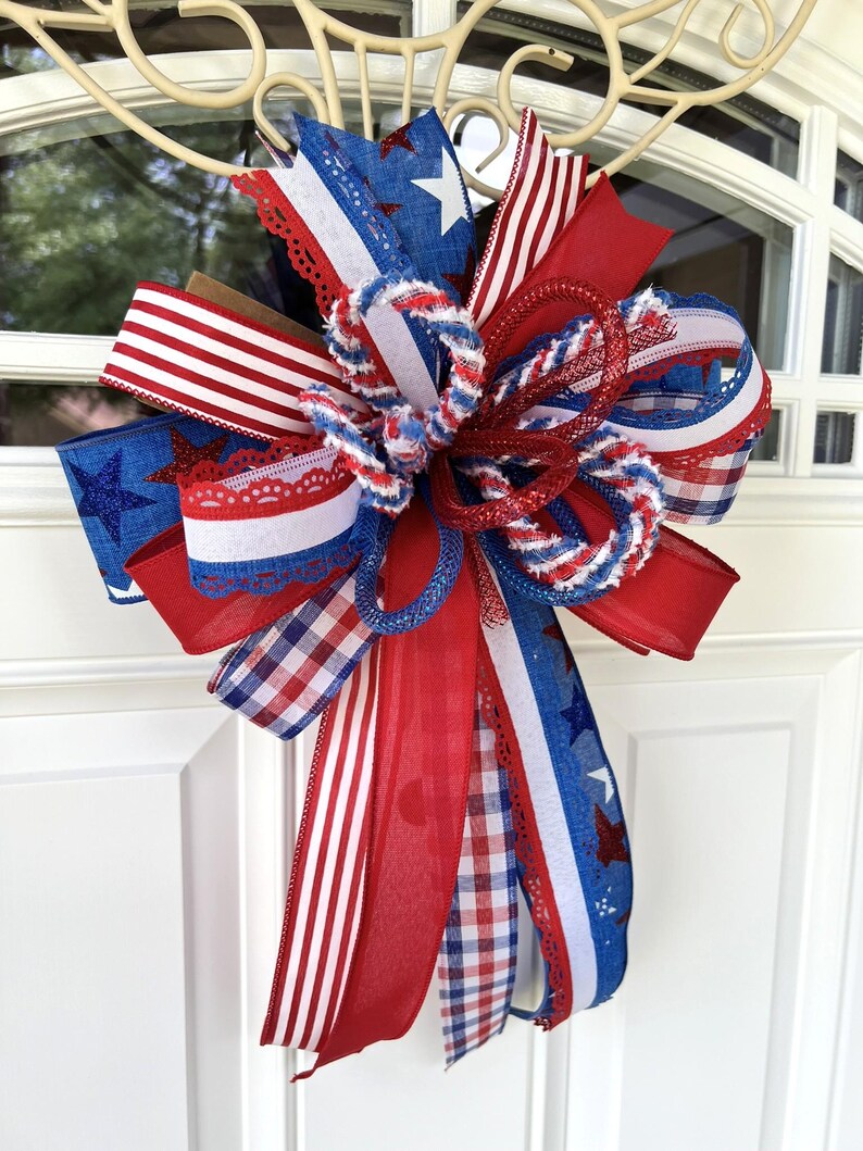 Op de afbeelding: Een decoratieve strik met een patriottisch thema in rood, wit en blauw. Het ontwerp bevat diverse patronen zoals sterren, strepen en gingham-ruitjes, gecombineerd met gestructureerd gaas en stoffen linten met een kanten rand.