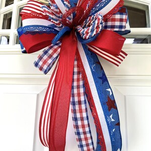 Op de afbeelding: Een decoratieve strik bestaande uit meerdere linten in rood, wit en blauw. Het ontwerp bevat patronen van sterren, strepen en vichy-ruitjes, waarbij sommige linten zijn voorzien van glitteraccenten en kanten randen, gebonden tot een grote strik.