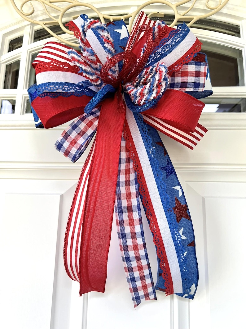 Op de afbeelding: Een decoratieve strik bestaande uit meerdere linten in rood, wit en blauw. Het ontwerp bevat patronen van sterren, strepen en vichy-ruitjes, waarbij sommige linten zijn voorzien van glitteraccenten en kanten randen, gebonden tot een grote strik.