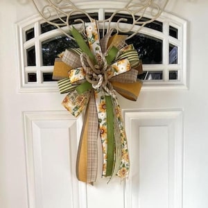 May include: A decorative bow made of multiple ribbons featuring sunflower patterns, solid olive green, and brown plaid textures. The bow has a central knot of natural jute rope and long, trailing ribbon tails hanging downward in a layered arrangement.