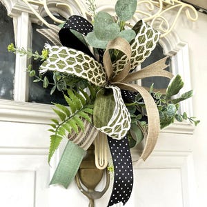 May include: A decorative door hanger featuring a multi-layered bow made of black and white polka dot ribbon, olive green linen ribbon, and a white lattice pattern ribbon, accented with artificial eucalyptus, fern leaves, and small yellow flowers.