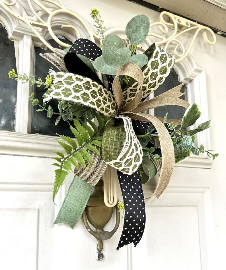 May include: A decorative door hanger featuring a multi-layered bow made of black and white polka dot ribbon, olive green linen ribbon, and a white lattice pattern ribbon, accented with artificial eucalyptus, fern leaves, and small yellow flowers.