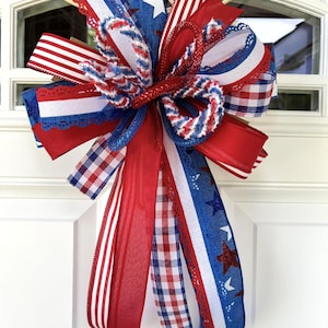 Op de afbeelding: Een decoratieve strik met een patriottisch thema, bestaande uit rode, witte en blauwe linten. Het ontwerp bevat diverse patronen zoals sterren, strepen en vichy-ruitjes, waarbij sommige linten glinsterende texturen en kantachtige randen hebben.