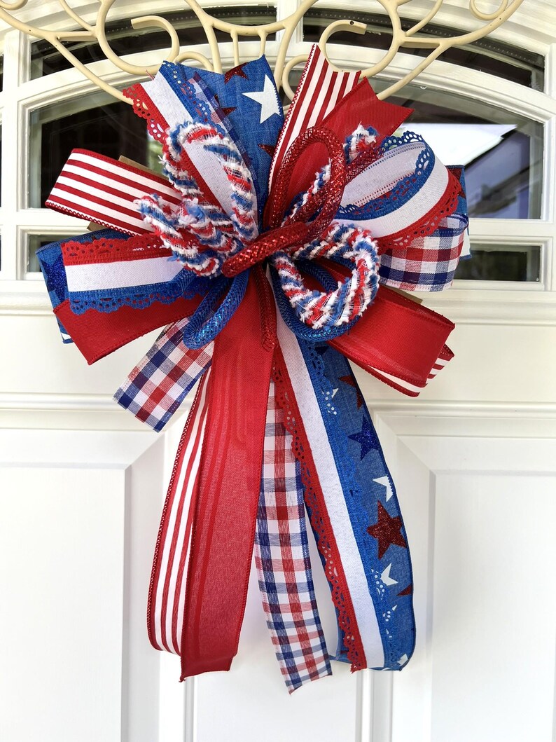 Op de afbeelding: Een decoratieve strik met een patriottisch thema, bestaande uit rode, witte en blauwe linten. Het ontwerp bevat diverse patronen zoals sterren, strepen en vichy-ruitjes, waarbij sommige linten glinsterende texturen en kantachtige randen hebben.