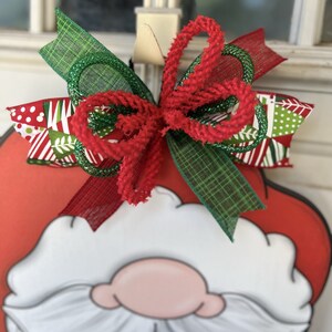 May include: A red and white Santa Claus door hanger with a green and red bow. The bow is made of plaid ribbon, red velvet ribbon, and green glitter ribbon.