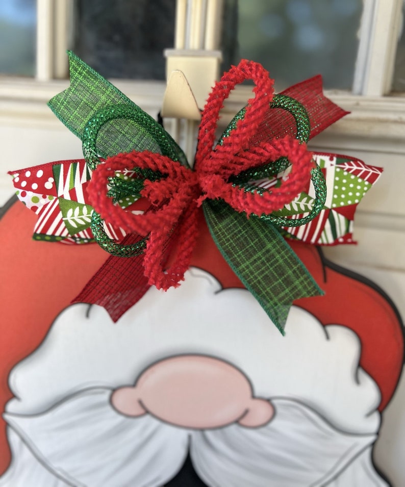 May include: A red and white Santa Claus door hanger with a green and red bow. The bow is made of plaid ribbon, red velvet ribbon, and green glitter ribbon.
