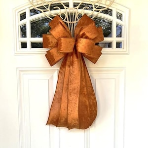 May include: A large, decorative bow made of shimmering, copper-colored fabric. The bow is attached to a white door with a decorative metal accent above. The bow's multiple loops and long, flowing tails create a festive and elegant look, suitable for seasonal decor.