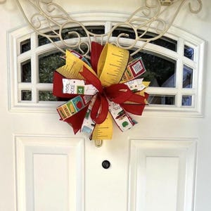 May include: A decorative bow featuring a school-themed pattern with yellow ribbons printed with ruler markings, solid red ribbons, and white ribbons decorated with colorful illustrations of apples, pencils, and the text School Rocks.