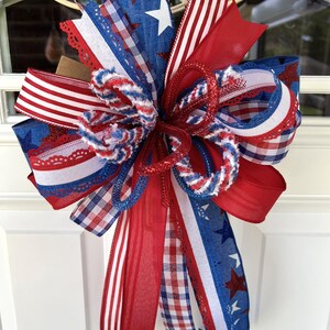 Op de afbeelding: Een decoratieve strik bestaande uit een mix van rode, witte en blauwe linten. Het ontwerp bevat patronen van sterren, strepen en gingham-ruitjes. De linten hebben gevarieerde texturen, waaronder transparante, effen en gerafelde randen.