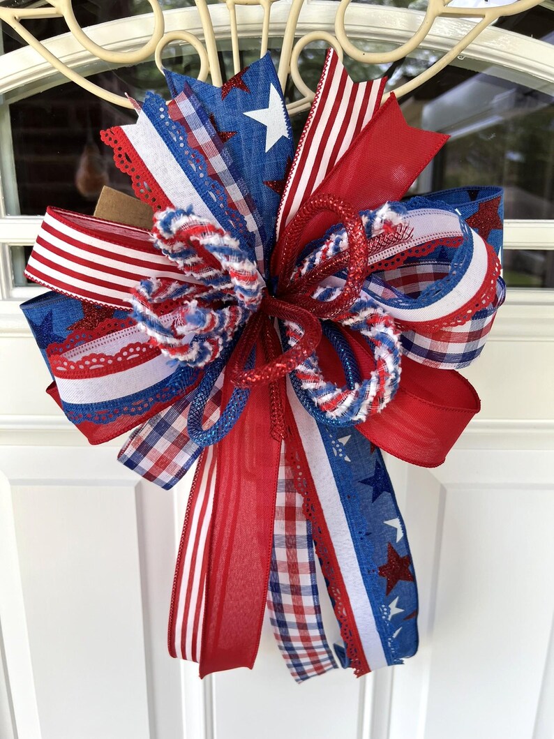 Op de afbeelding: Een decoratieve strik bestaande uit een mix van rode, witte en blauwe linten. Het ontwerp bevat patronen van sterren, strepen en gingham-ruitjes. De linten hebben gevarieerde texturen, waaronder transparante, effen en gerafelde randen.