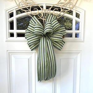 May include: A decorative bow made from fabric ribbon featuring a pattern of vertical stripes in shades of forest green, olive green, and white. The bow has a large, multi-looped top section and two long, flowing tails hanging down from the center.