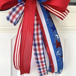 Op de afbeelding: Een decoratieve strik gemaakt van meerdere linten in de kleuren rood, wit en blauw. De linten bevatten patronen van rood-witte strepen, rood-blauwe vichy-ruitjes, effen rood en een blauw lint met witte en rode glinsterende sterren.