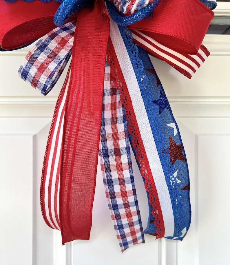 Op de afbeelding: Een decoratieve strik gemaakt van meerdere linten in de kleuren rood, wit en blauw. De linten bevatten patronen van rood-witte strepen, rood-blauwe vichy-ruitjes, effen rood en een blauw lint met witte en rode glinsterende sterren.