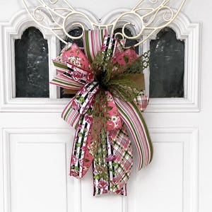 May include: A decorative bow featuring a mix of patterned ribbons in pink, green, and black. The bow includes floral, striped, and plaid designs, accented with faux greenery. The bow is attached to a white, ornate metal wreath frame, suitable for door decor.
