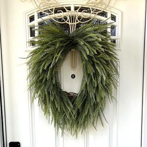 May include: A large, circular green fern wreath hangs on a white door. The wreath is made of faux fern leaves and has a natural, rustic appearance. A decorative metal piece is above the door. The door has a window with a decorative design.