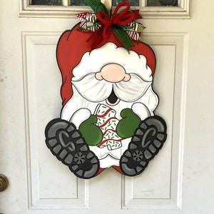 May include: A Christmas door hanger featuring a cartoon Santa Claus wearing a red hat, white beard, and green mittens. He is holding a candy cane and has a big smile on his face. The hanger is decorated with a red and green bow.