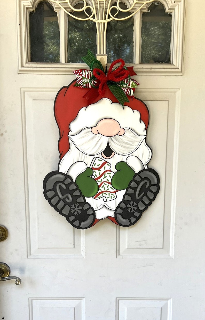 May include: A Christmas door hanger featuring a cartoon Santa Claus wearing a red hat, white beard, and green mittens. He is holding a candy cane and has a big smile on his face. The hanger is decorated with a red and green bow.