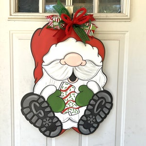 May include: A wooden Christmas door hanger featuring a Santa Claus gnome holding a wrapped gift. The gnome is wearing a red hat, white beard, and green mittens. The gift is wrapped in red and white paper with green sprinkles.