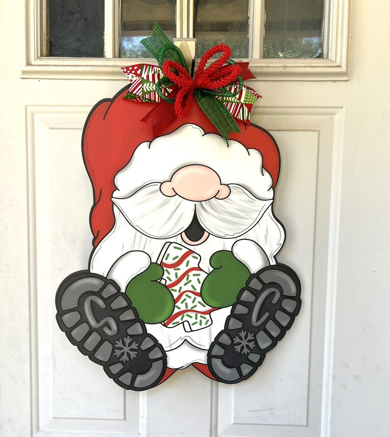 May include: A wooden Christmas door hanger featuring a Santa Claus gnome holding a wrapped gift. The gnome is wearing a red hat, white beard, and green mittens. The gift is wrapped in red and white paper with green sprinkles.