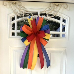 May include: A colorful rainbow-themed bow made of various ribbons, attached to a white door. The bow features a vibrant array of colors, including red, orange, yellow, green, blue, and purple. The bow is a decorative accent for the door.