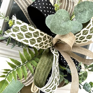 May include: A decorative bow featuring a mix of ribbons including black with white polka dots, olive green with a white lattice pattern, tan linen, and striped fabric. The bow is accented with artificial green eucalyptus and fern foliage.