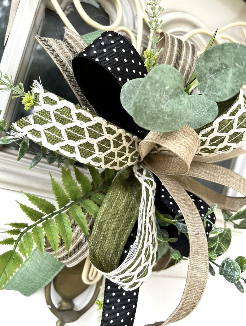 May include: A decorative bow featuring a mix of ribbons including black with white polka dots, olive green with a white lattice pattern, tan linen, and striped fabric. The bow is accented with artificial green eucalyptus and fern foliage.