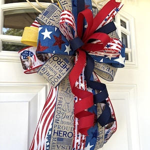 May include: A patriotic door bow featuring red, white, and blue ribbons with star and stripe patterns. The bow includes ribbons with text such as "Duty, Honor, Proud, Hero" and "USA Marines". The bow is attached to a white door.