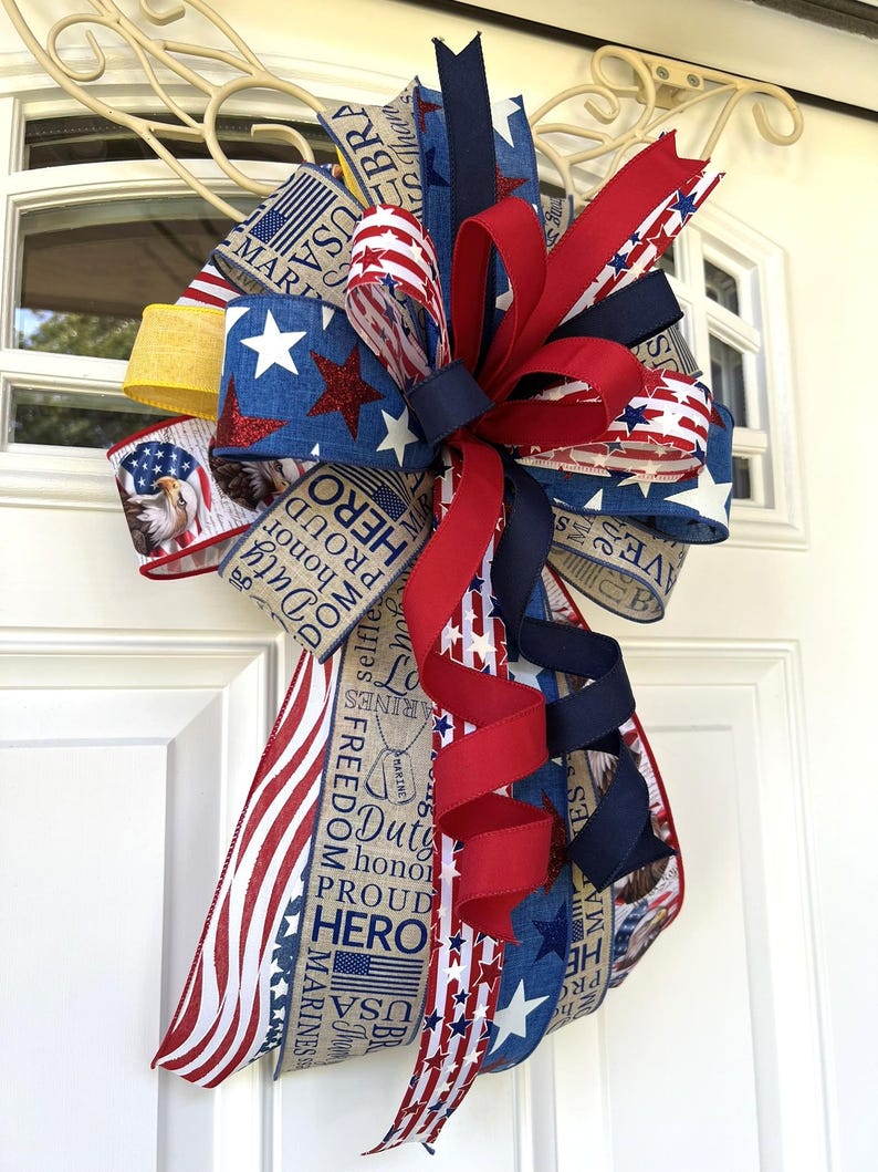 May include: A patriotic door bow featuring red, white, and blue ribbons with star and stripe patterns. The bow includes ribbons with text such as "Duty, Honor, Proud, Hero" and "USA Marines". The bow is attached to a white door.