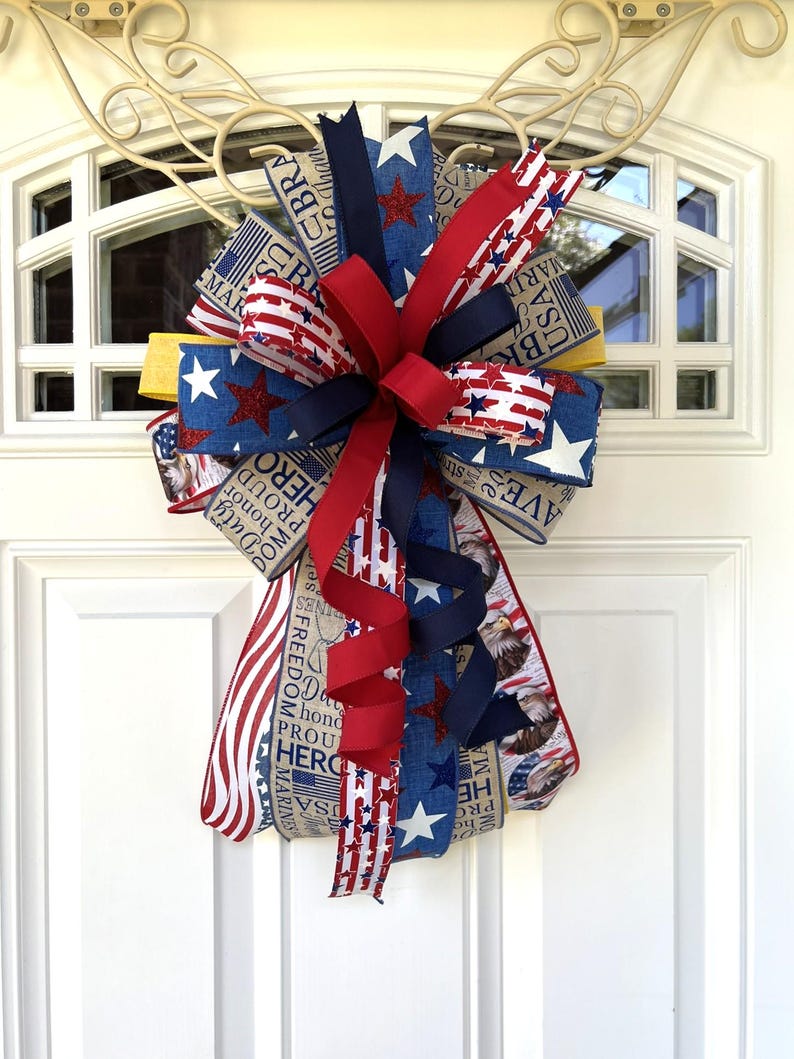 May include: A patriotic door bow featuring red, white, and blue ribbons with star and stripe patterns. The bow includes ribbons with text such as "USA", "Freedom", and "Hero". A decorative accent for Independence Day or other patriotic holidays.