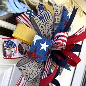 May include: A patriotic bow featuring red, white, and blue ribbons with stars, stripes, and eagle designs. Some ribbons have text that reads "USA Marines" and "Duty, Honor, Proud". This decorative bow is perfect for Independence Day or other patriotic holidays.