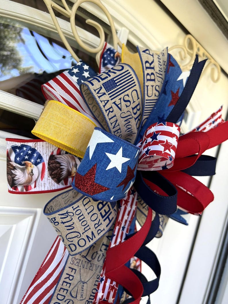May include: A patriotic bow featuring red, white, and blue ribbons with stars, stripes, and eagle designs. Some ribbons have text that reads "USA Marines" and "Duty, Honor, Proud". This decorative bow is perfect for Independence Day or other patriotic holidays.