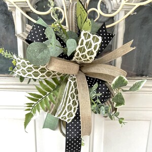 May include: A decorative bow arrangement featuring black ribbon with white polka dots, tan burlap ribbon, and a green ribbon with a white lattice pattern. The piece is accented with artificial fern fronds, eucalyptus leaves, and a green butterfly.