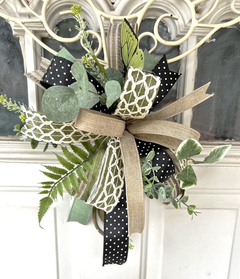 May include: A decorative bow arrangement featuring black ribbon with white polka dots, tan burlap ribbon, and a green ribbon with a white lattice pattern. The piece is accented with artificial fern fronds, eucalyptus leaves, and a green butterfly.