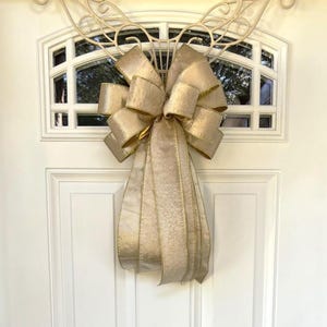 May include: A large, gold-colored bow with long, flowing ribbons hangs on a white door. The bow is made of a shimmering fabric with gold trim. The door has a decorative window and a white metal accent above the bow.
