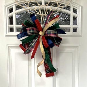 May include: A decorative bow with a mix of red, green, navy, and gold ribbons. The bow features a plaid pattern and velvet textures, perfect for holiday decor. It is attached to a white door with a decorative metal accent.