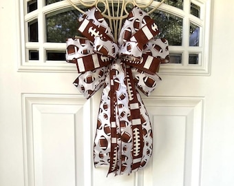 May include: A decorative bow made from white ribbon featuring a repeating pattern of brown American footballs and brown textured ribbon strips. The bow has multiple loops and two long, hanging tails, showcasing a sports-themed design.