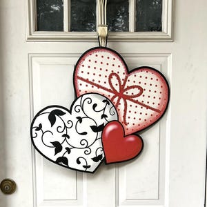 May include: Three heart-shaped decorations for a door. The largest heart is white with red polka dots and a red bow. The middle heart is white with black floral designs. The smallest heart is red.