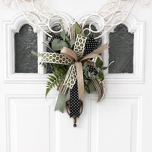 May include: A decorative door wreath featuring a mix of ribbons in black with white polka dots, brown, and green. The wreath includes greenery and is attached to a white door with a decorative metal accent.