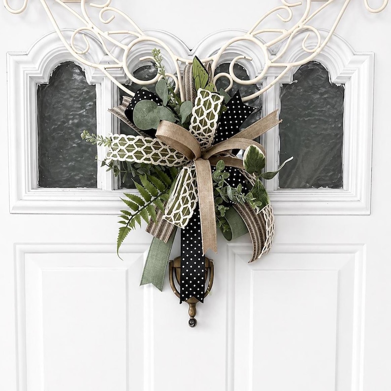 May include: A decorative door swag featuring a mix of ribbons in black with white polka dots, solid tan, and green with a white lattice pattern. The arrangement includes artificial green fern leaves and eucalyptus sprigs attached to a metal door knocker.