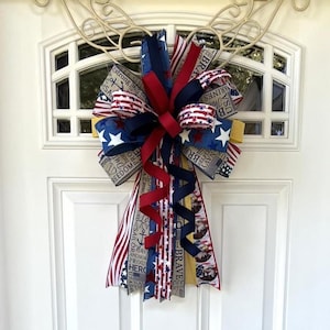 May include: A patriotic door bow featuring red, white, and blue ribbons. The bow includes ribbons with star and stripe patterns, along with text that reads "Brave," "Freedom," and "Anchor." The bow is designed to be hung on a door.