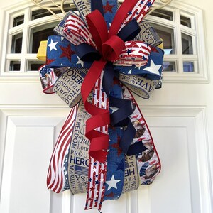 May include: A patriotic bow featuring red, white, and blue ribbons with star and stripe patterns. The bow includes ribbons with text such as "Freedom", "Hero", and "Marines". The bow is attached to a white door with a decorative metal accent.