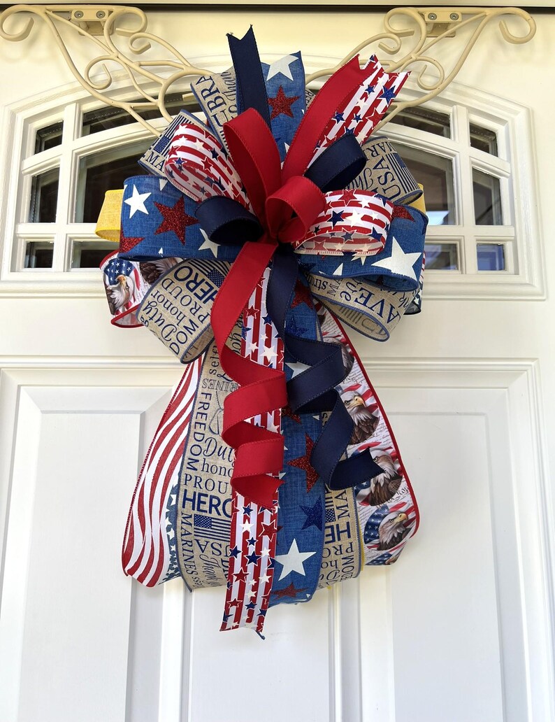 May include: A patriotic bow featuring red, white, and blue ribbons with star and stripe patterns. The bow includes ribbons with text such as "Freedom", "Hero", and "Marines". The bow is attached to a white door with a decorative metal accent.