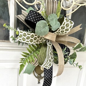 May include: A decorative bow featuring a mix of black ribbon with white polka dots, solid tan burlap, and olive green ribbon with a white lattice pattern. The arrangement is accented with artificial green eucalyptus leaves and fern sprigs.