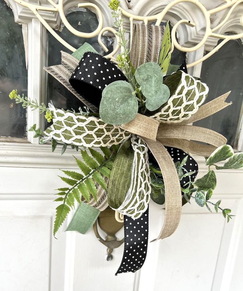 May include: A decorative bow featuring a mix of black ribbon with white polka dots, solid tan burlap, and olive green ribbon with a white lattice pattern. The arrangement is accented with artificial green eucalyptus leaves and fern sprigs.