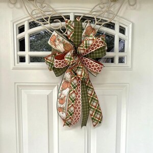 May include: A decorative fall-themed bow with multiple ribbons in various patterns and colors. The bow features ribbons with pumpkin and plaid designs in orange, green, and brown hues. It is attached to a white door with a decorative window.