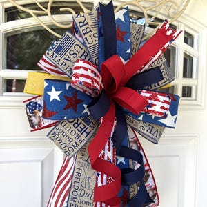 May include: A patriotic door bow featuring red, white, and blue ribbons with star and stripe patterns. The bow includes ribbons with text such as "Freedom", "Hero", and "Marine". A decorative accent for patriotic holidays.