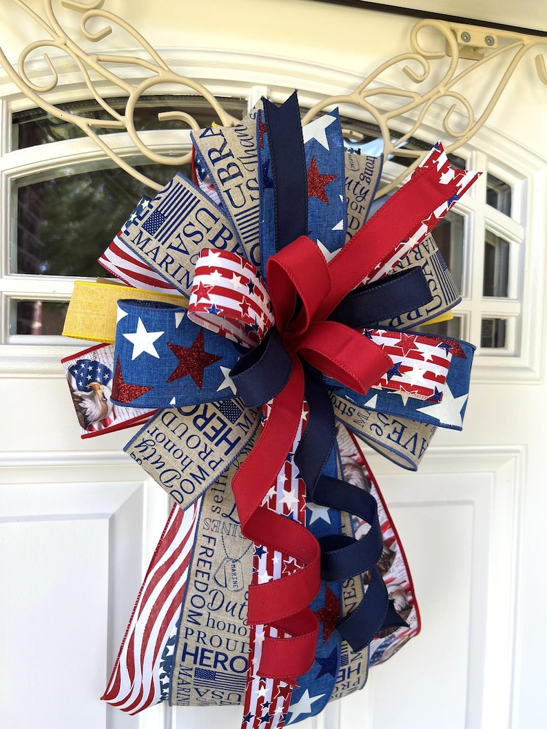 May include: A patriotic door bow featuring red, white, and blue ribbons with star and stripe patterns. The bow includes ribbons with text such as "Freedom", "Hero", and "Marine". A decorative accent for patriotic holidays.