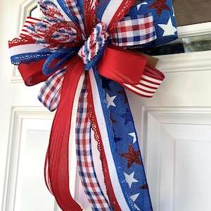 Op de afbeelding: Een decoratieve strik met een patriottisch thema, bestaande uit rode, witte en blauwe linten. Het ontwerp bevat effen rode linten, blauwe linten met witte en rode glittersterren, en rood-wit-blauwe ruit- en streepmotieven met kanten randen.