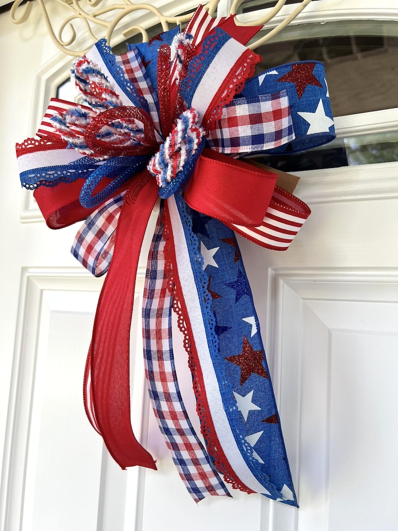 Op de afbeelding: Een decoratieve strik met een patriottisch thema, bestaande uit rode, witte en blauwe linten. Het ontwerp bevat effen rode linten, blauwe linten met witte en rode glittersterren, en rood-wit-blauwe ruit- en streepmotieven met kanten randen.